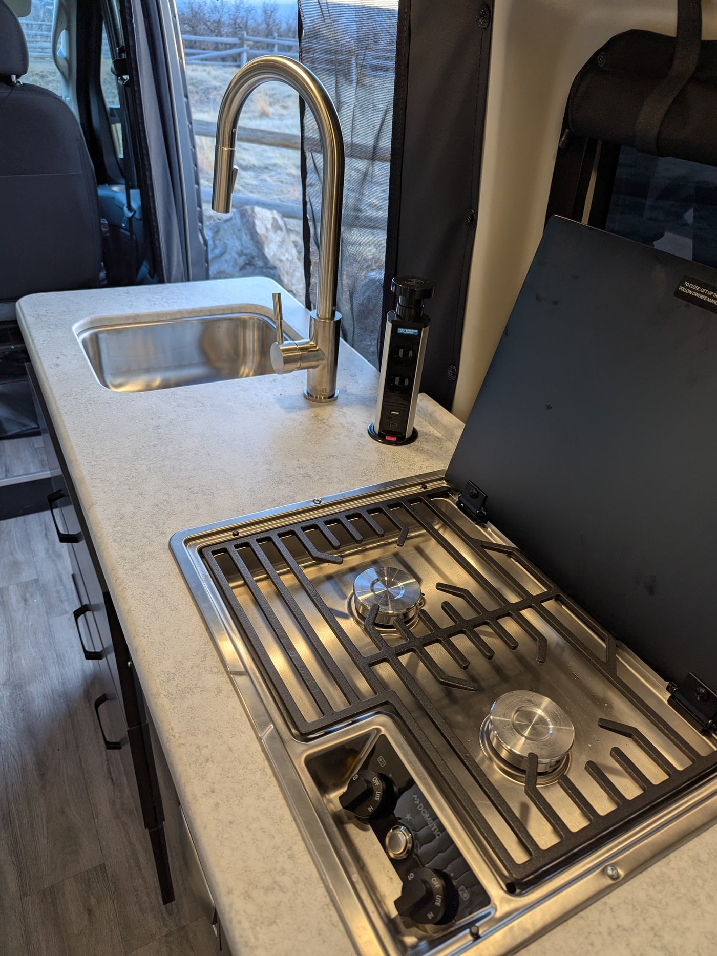 Inside an RV rental, a compact kitchenette features a stainless steel sink, modern faucet, Dometic stove, and pop-up power outlet. — photo 10