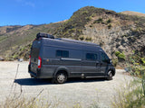 A dark grey Winnebago Solis camper van, available as an **rv rental**, parked on a gravel road with scenic mountains.