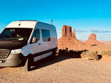 An RV rental van, white with a black hood, is parked on a gravel lot before majestic red rock formations under a blue sky.
