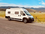 An RV rental - a white Winnebago Solis camper van - parked on a gravel road, with mountains & fields in the background.