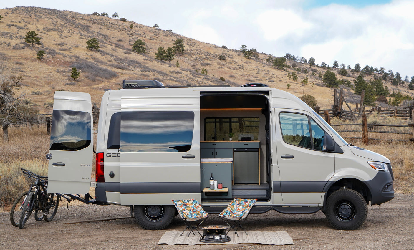 This light gray camper van (rv rental) with open doors, bikes, chairs, and a grill is set up for camping in a scenic, arid landscape.