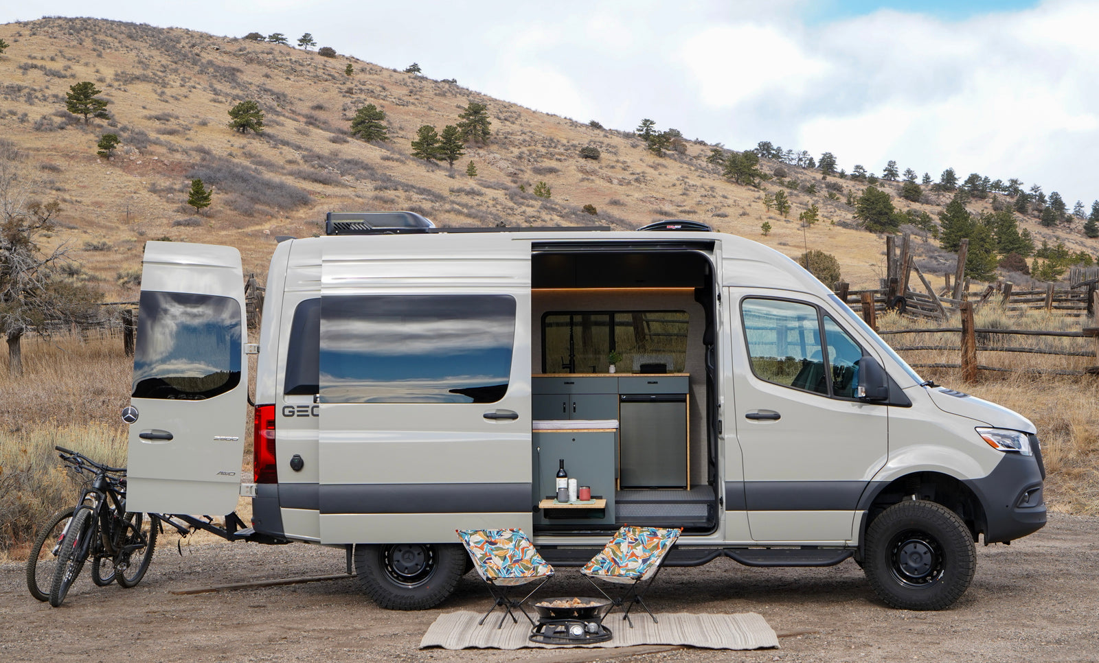 This light gray camper van (rv rental) with open doors, bikes, chairs, and a grill is set up for camping in a scenic, arid landscape.