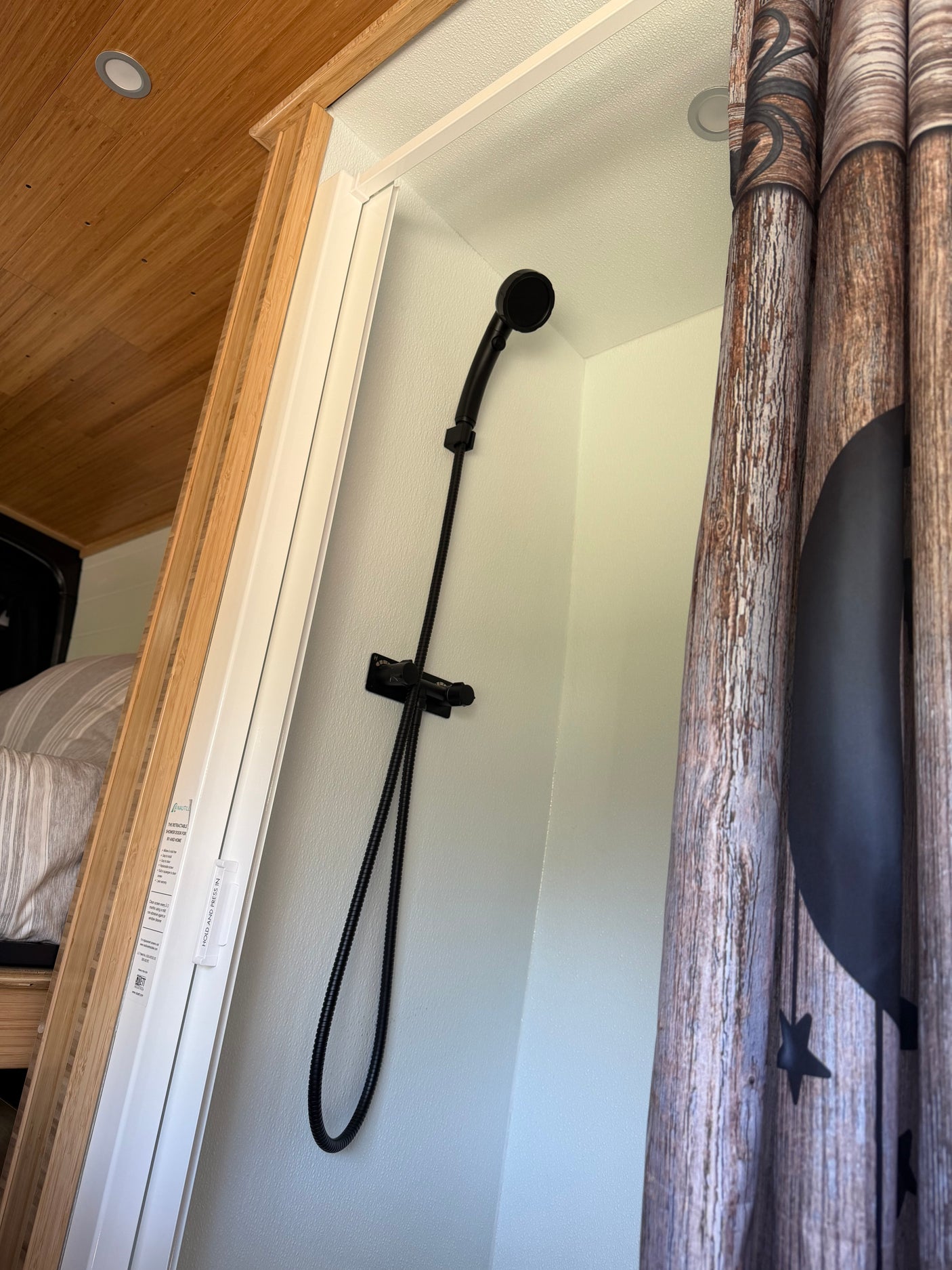 Compact RV rental shower with black handheld showerhead, wood-look curtain, and space-saving retractable door.