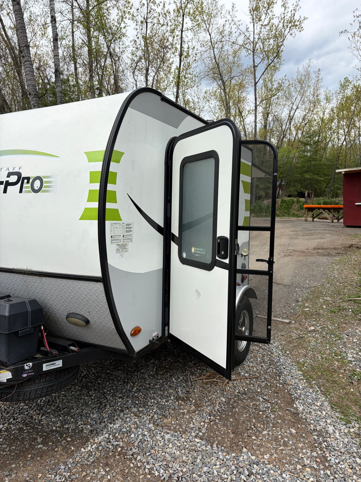 White and green travel trailer RV rental with door open, revealing screen. Parked on gravel, surrounded by trees for camping. — photo 2