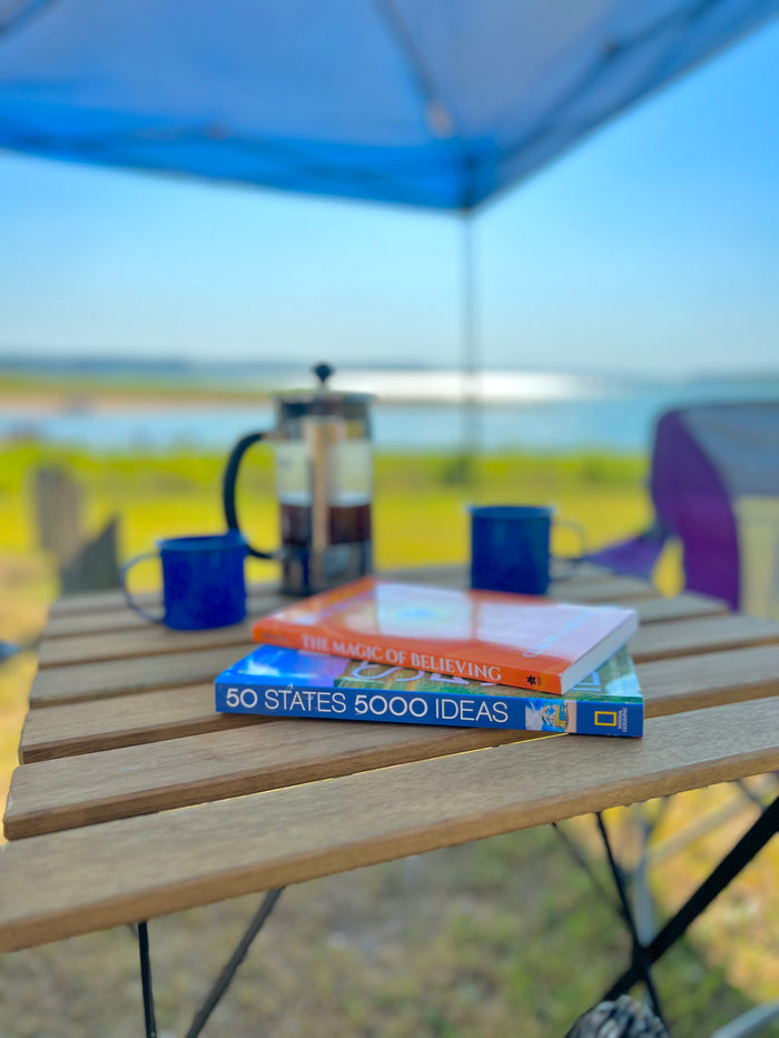 RV rental escape: Books, coffee, and peaceful lake views under a blue canopy on a wooden table.