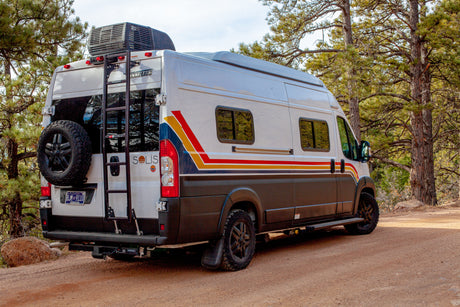 This white Winnebago Solis rv rental, with a spare tire and ladder, rests on a dirt road among pines.