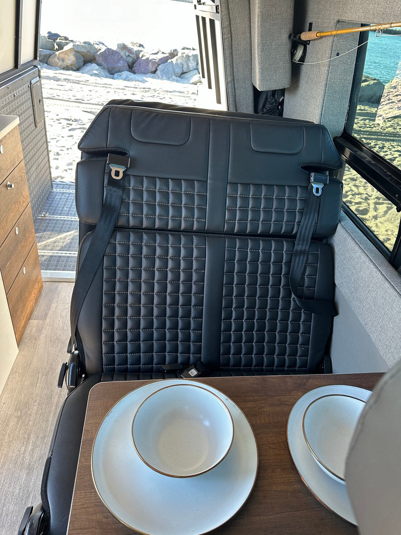 RV rental interior: black seating with seatbelts, wooden dining table with place settings, fishing rod mounted, and a sandy beach view outside. — photo 5