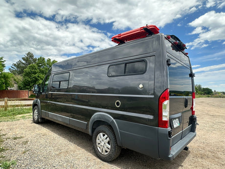 Dark gray camper van, equipped with roof recovery boards & rear ladder, ideal for RV rental adventures. Parked on gravel.