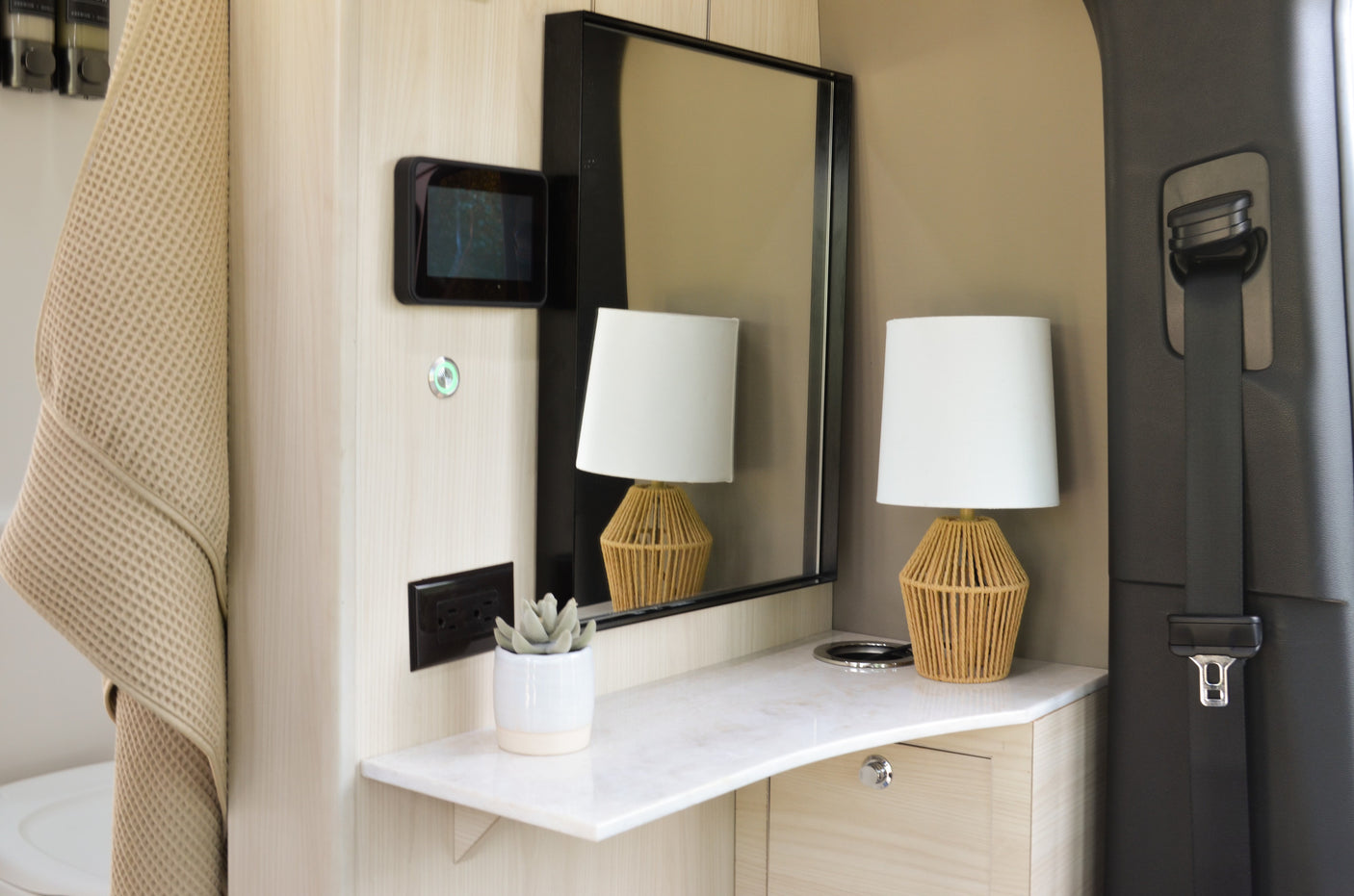 Modern RV rental interior showing a light wood vanity, mirror, woven lamp, and wall-mounted screen. Seatbelt visible.
