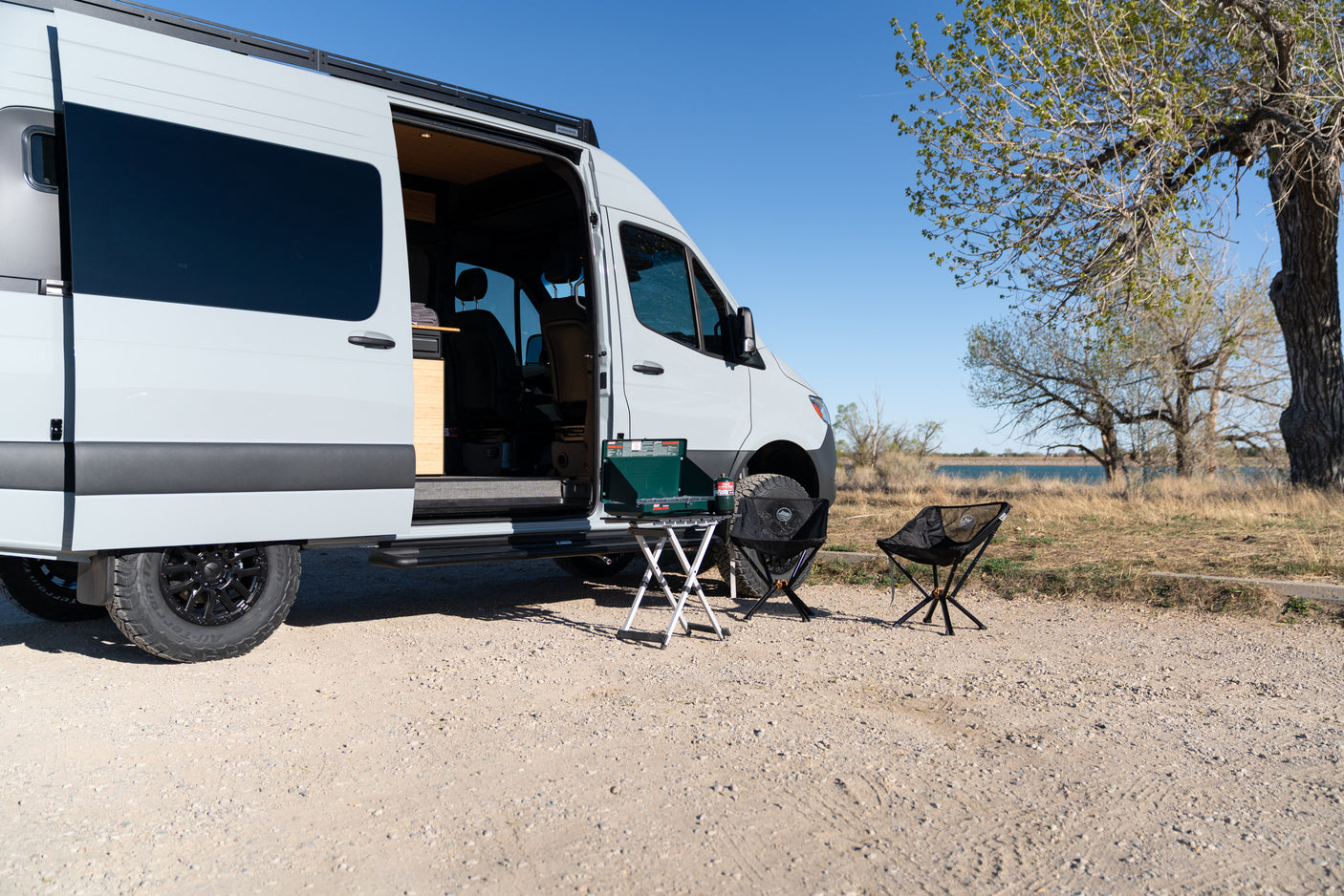 This RV rental camper van setup features an open door, camp stove, and chairs by a scenic lake, perfect for outdoor adventures.