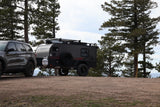Rugged dark SUV tows an off-road 'Flyer' camper, ideal for an rv rental adventure among tall pines.
