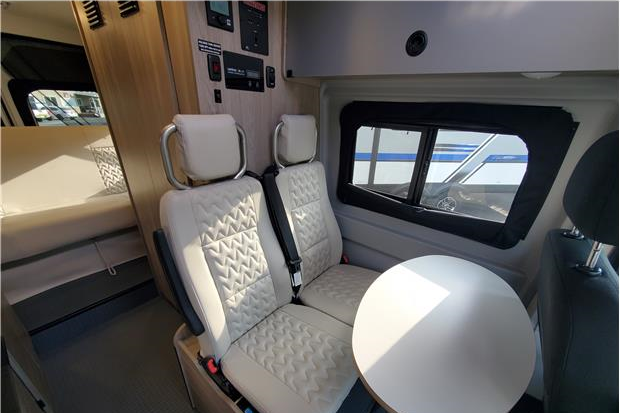 Experience an **RV rental**! Modern camper van interior features two quilted seats, a table, and a rear bed area.