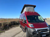 A red pop-top camper van, ideal for rv rental, parked by a fence with industrial cranes in the background.