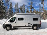 A sleek RV rental, a silver camper van, stands amidst a snowy forest with pine trees under blue skies.