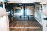 Organized under-bed storage in an RV rental camper van, featuring modular panels, LED lighting, and tie-downs for travel gear.