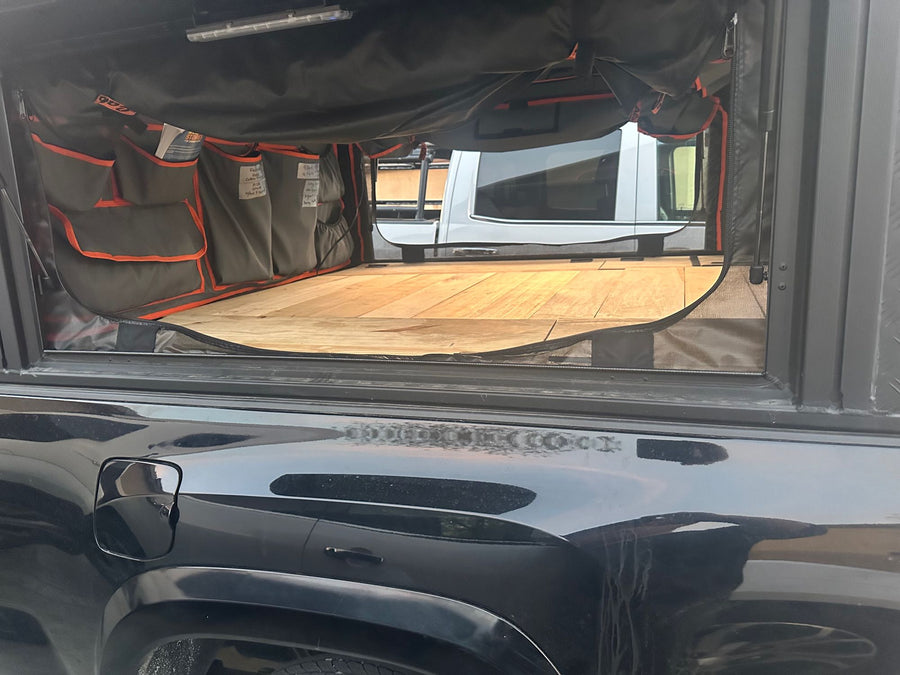  and front seats. Excellent for your next RV rental journey.|Modified black truck with rooftop tent & awning. An RV rental is visible by mountains & water in a gravel campground.|Optimized RV rental cargo space featuring wooden benches