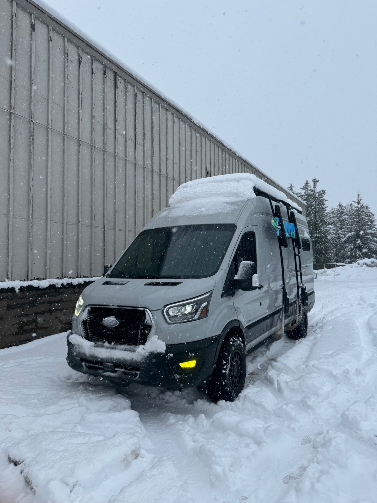 This RV rental is ready for winter adventure! A grey camper van covered in snow, with falling snowflakes, parked by a building. — photo 3