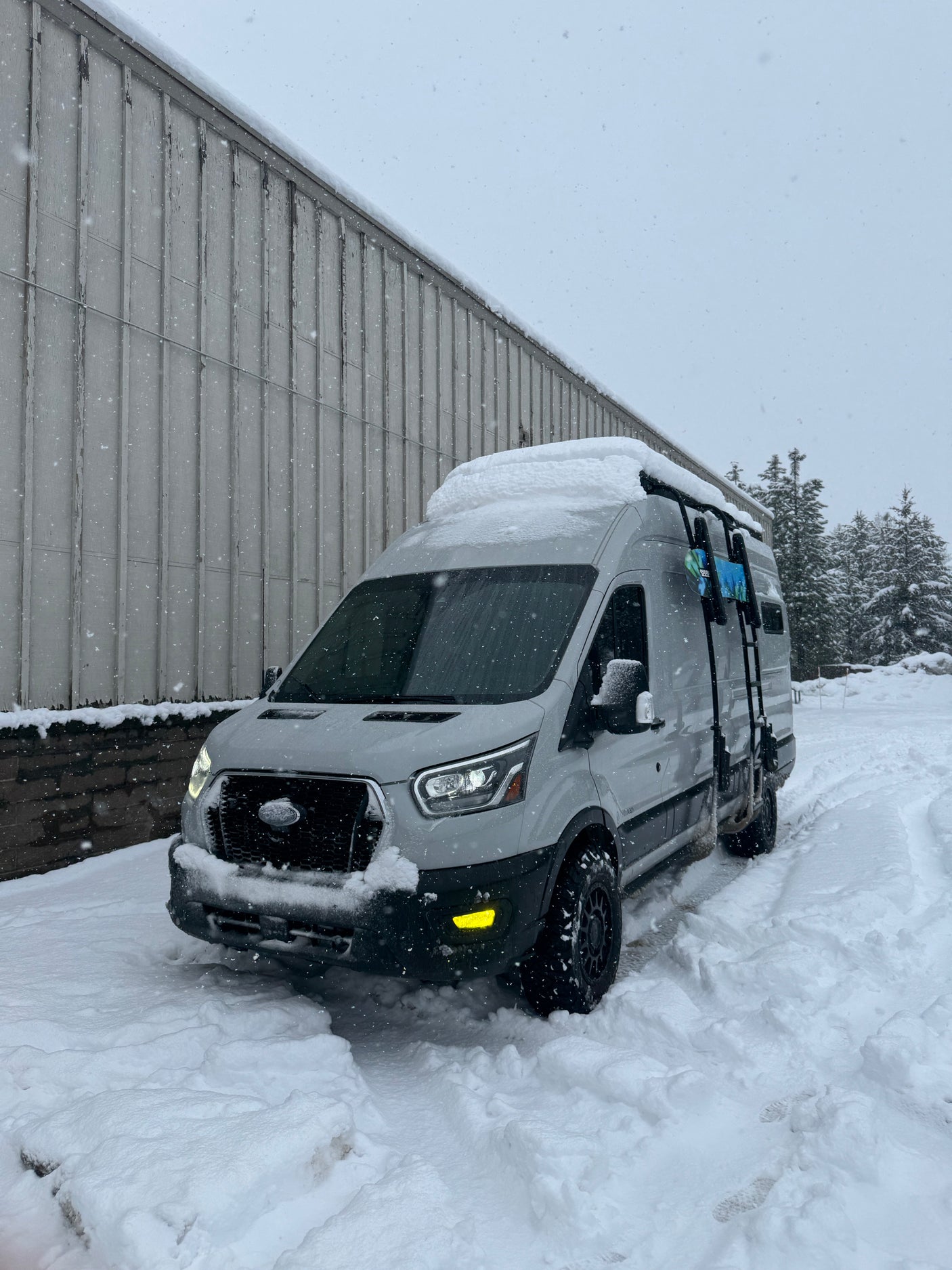 This RV rental is ready for winter adventure! A grey camper van covered in snow, with falling snowflakes, parked by a building.