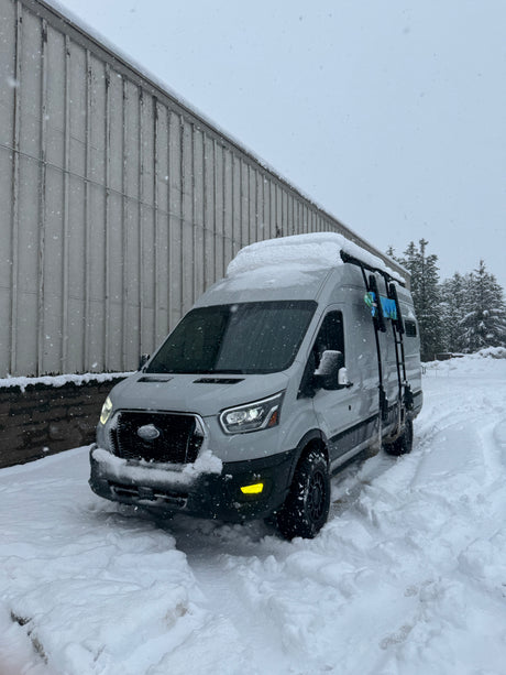 This RV rental is ready for winter adventure! A grey camper van covered in snow, with falling snowflakes, parked by a building.