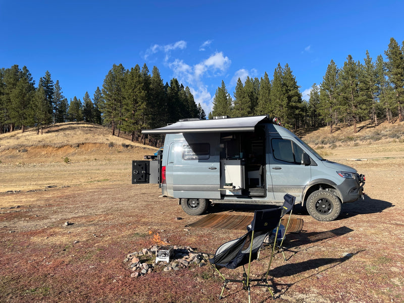 Sunny RV rental campsite: A grey camper van, awning out, alongside a campfire & chairs in a forest clearing for camping. — photo 6