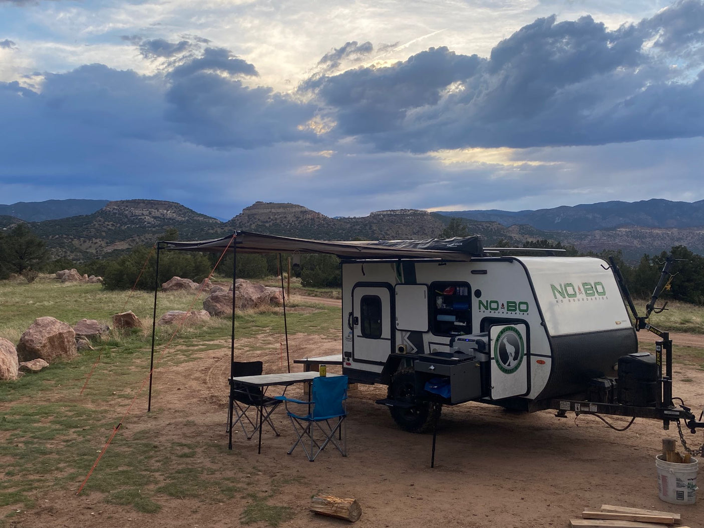 Scenic RV rental travel trailer with an extended awning, table, and chairs set up at a mountain campsite under cloudy skies.