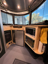 This RV rental interior features a microwave, refrigerator, and clever storage. Expansive windows reveal the sky and trees.