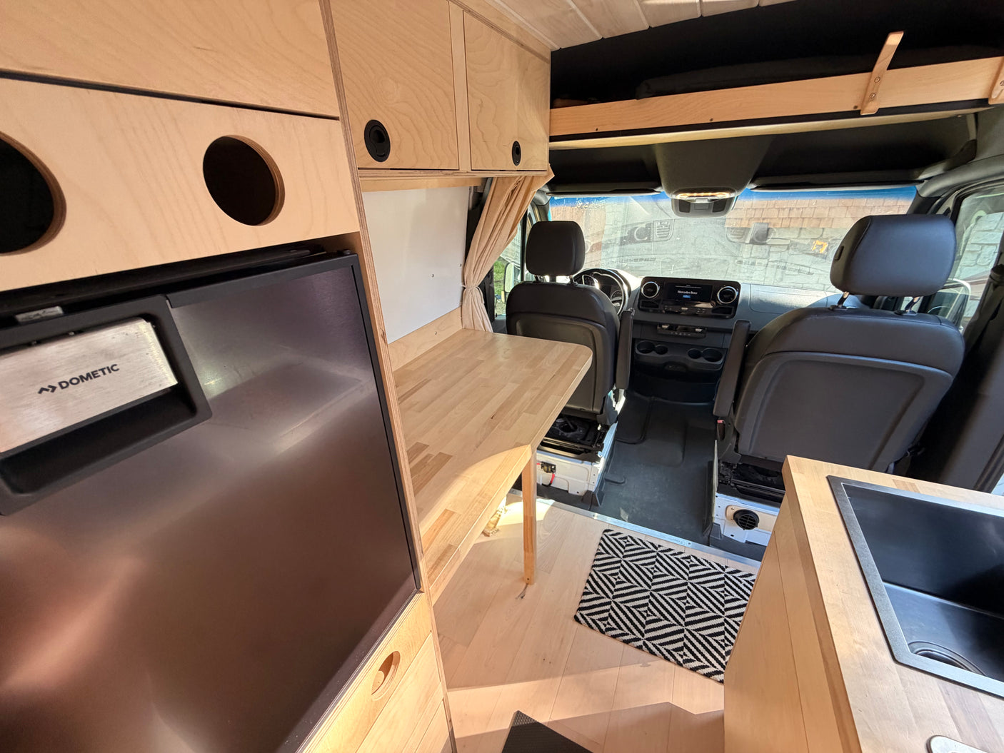 Inside an RV rental conversion van: light wood cabinets, Dometic fridge, sink, and front seats for a cozy camper experience.
