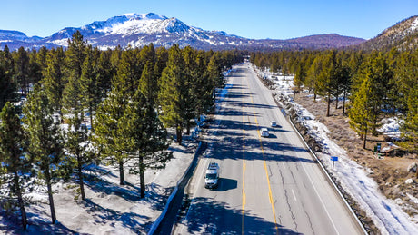 White RV rental on a scenic highway through snowy pine forests with majestic mountains in the background.