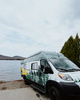 An rv rental camper van with mountain decals is parked by a scenic lake with hills, ready for adventure.