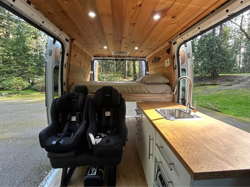  snow-capped mountains with a river & lush green forest.|Family-friendly RV rental interior: Cozy wood-paneled camper van with kitchen — photo 1