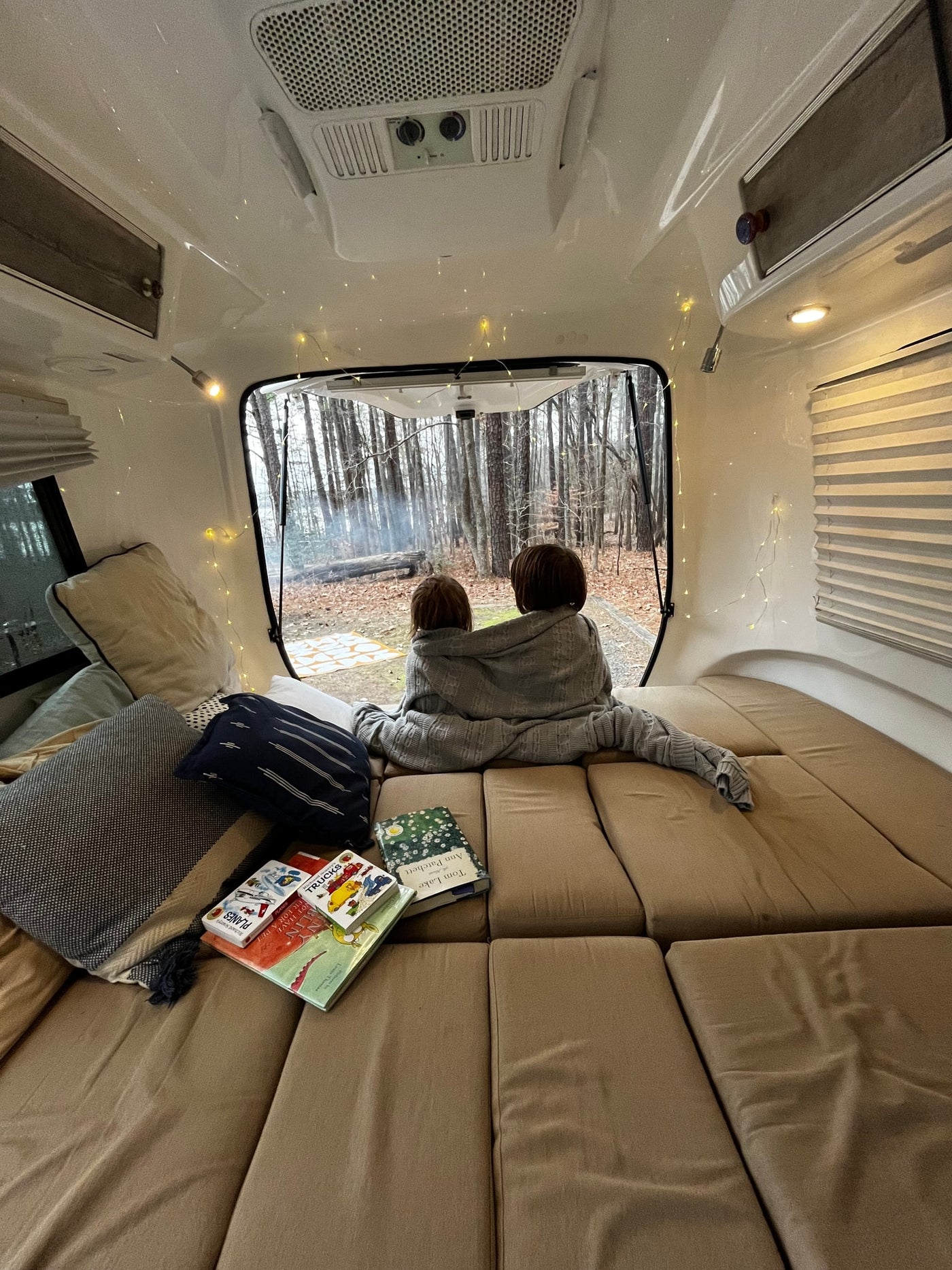 Inside a cozy **rv rental**: two children under a blanket watch a forest campfire. Books and string lights add to the warm atmosphere. — photo 6