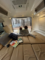 Inside a cozy **rv rental**: two children under a blanket watch a forest campfire. Books and string lights add to the warm atmosphere.