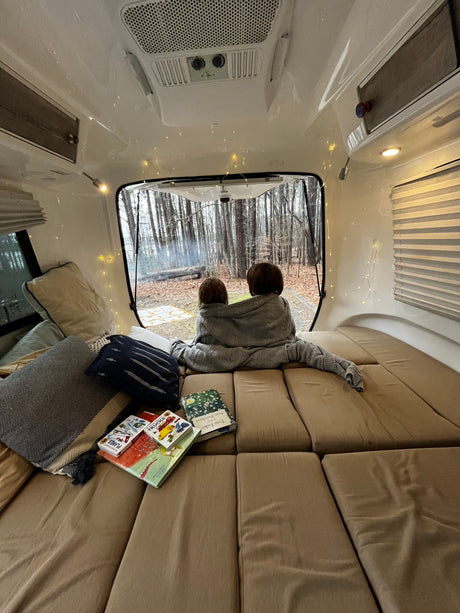 Inside a cozy **rv rental**: two children under a blanket watch a forest campfire. Books and string lights add to the warm atmosphere.