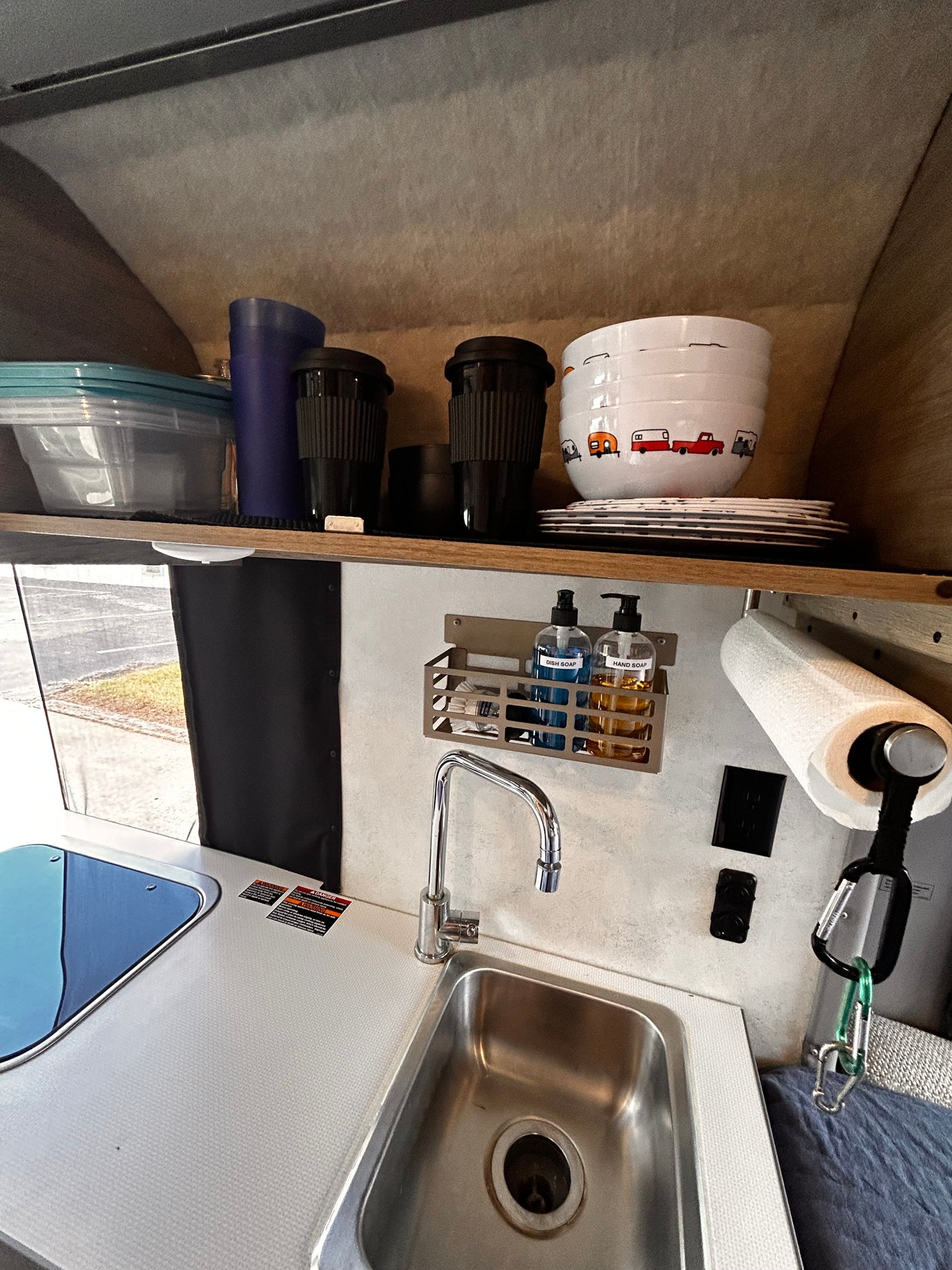 Inside an rv rental: a functional kitchenette with sink, covered stovetop, dishes, and cleaning supplies on shelves.