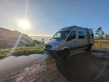 Scenic RV rental: Silver camper van by the ocean at sunset, parked on a dirt path next to a reflective puddle.