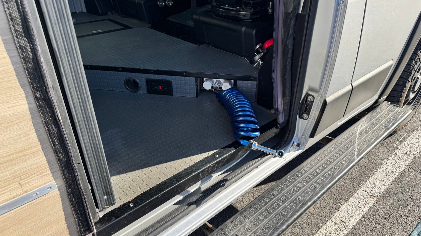 An RV rental van's open side door reveals a utility panel with quick-connect water fittings & a blue coiled wash hose.