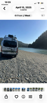 Scenic rv rental adventure: A white Winnebago Revel camper van parked on a rocky riverbank with forested hills and a calm river.