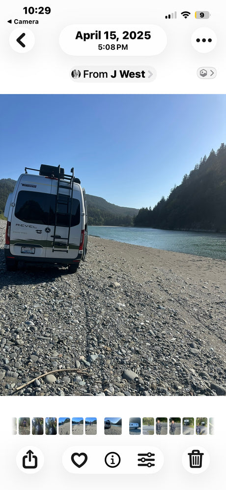 Scenic rv rental adventure: A white Winnebago Revel camper van parked on a rocky riverbank with forested hills and a calm river.