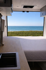 RV rental with ocean views! Inside a camper van, a bed on the right and a kitchen with a sink on the left face the sea.
