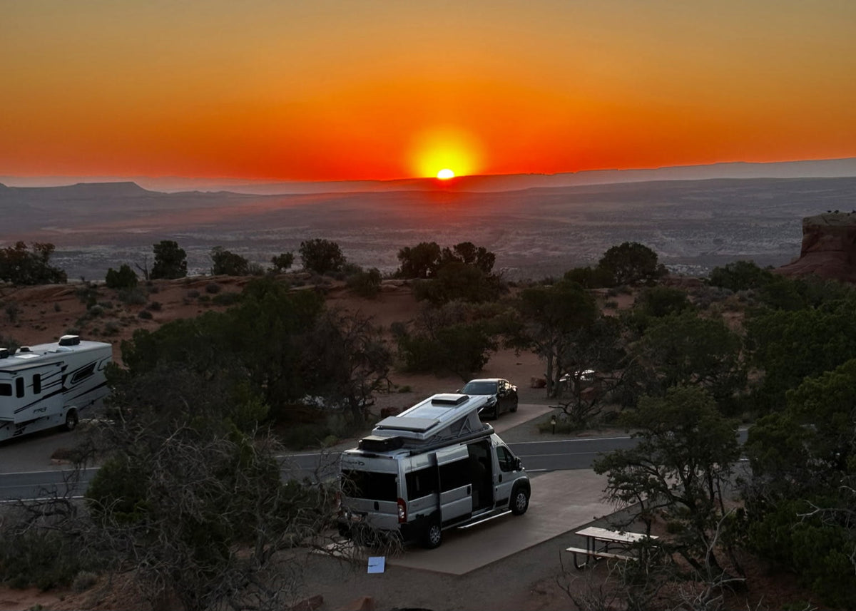 Experience adventure! An RV rental and another RV are parked at a desert campsite under a stunning sunset.|Vibrant sunset at an RV rental campsite. A silver RV is parked near a crackling campfire
