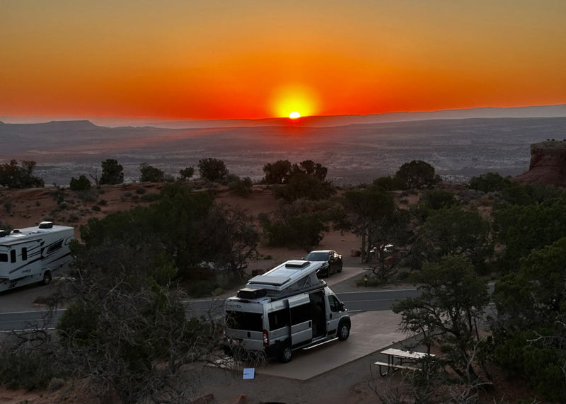 Experience adventure! An RV rental and another RV are parked at a desert campsite under a stunning sunset.|Vibrant sunset at an RV rental campsite. A silver RV is parked near a crackling campfire — photo 1