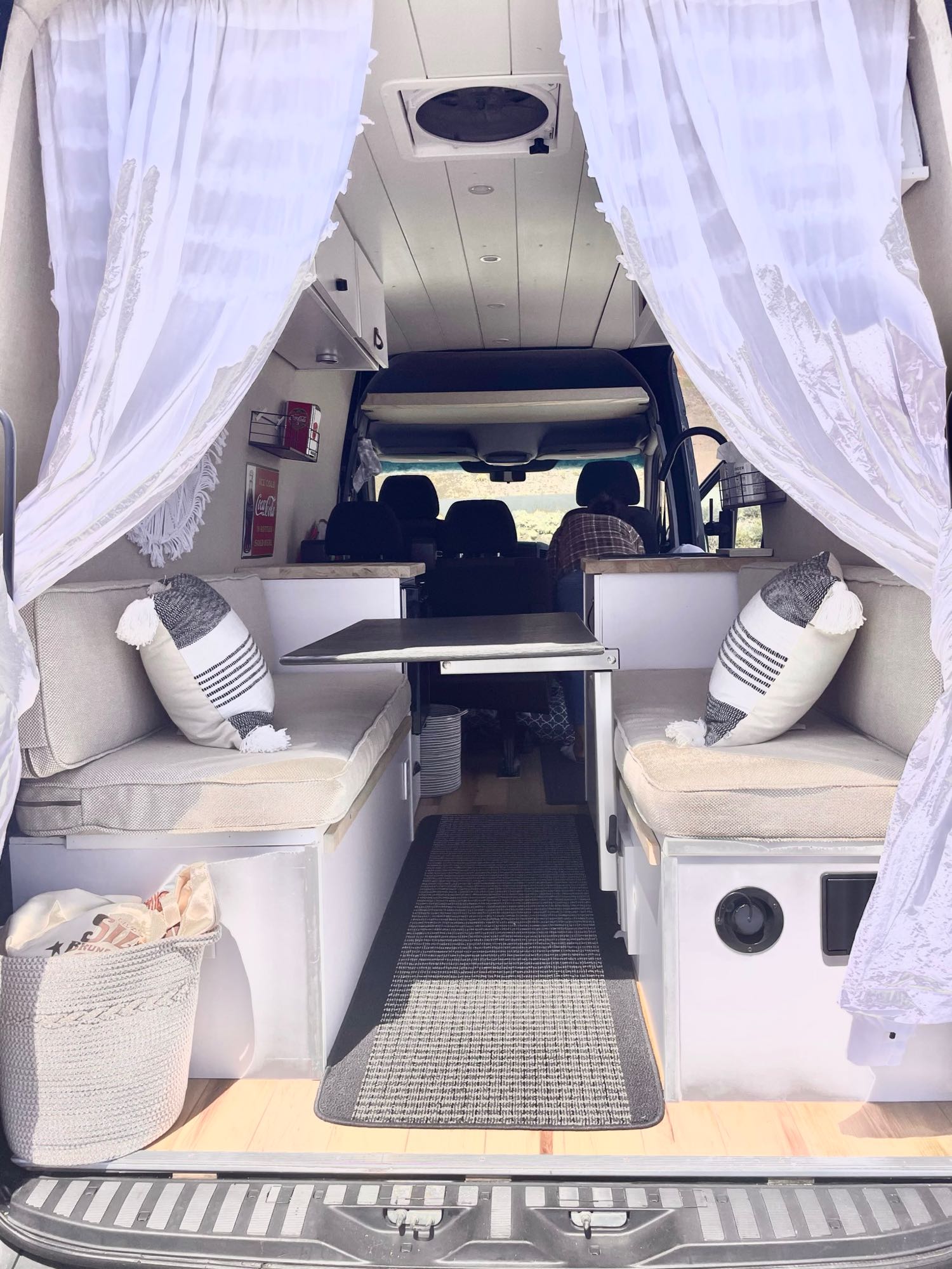 Cozy RV rental interior shows a dinette with benches, table, white curtains. A person is visible in the driver's seat.