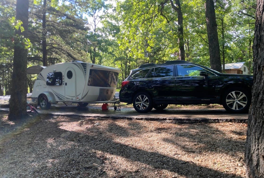 A white teardrop RV rental camper is hitched to a black Subaru Outback SUV, parked in a shady wooded campground.