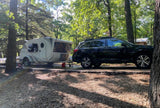 A white teardrop RV rental camper is hitched to a black Subaru Outback SUV, parked in a shady wooded campground.