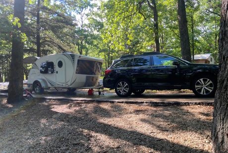 A white teardrop RV rental camper is hitched to a black Subaru Outback SUV, parked in a shady wooded campground.