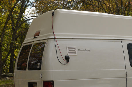 White Belaire RV rental parked amongst trees, showing its high roof, solar wiring connection, and rear windows.