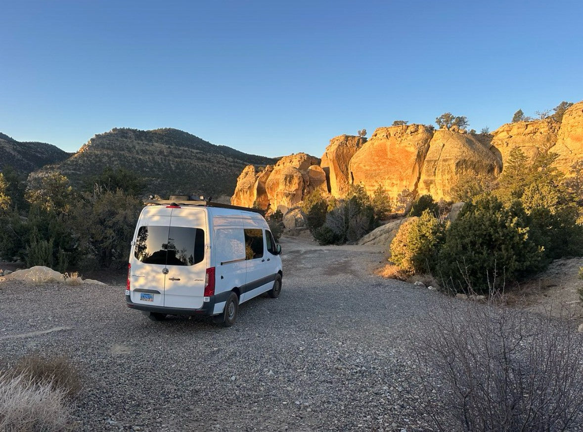 A white camper van, ideal for RV rental, on a gravel road by sunlit orange sandstone cliffs in a desert setting.