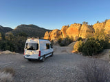A white camper van, ideal for RV rental, on a gravel road by sunlit orange sandstone cliffs in a desert setting.