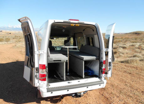White camper van RV rental with open back, revealing comfortable interior: convertible beds, table. Parked in desert.