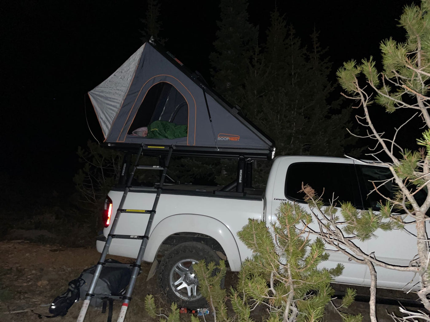 Nighttime RV rental: a white truck with an open rooftop tent, ladder, and sleeping bag visible, surrounded by dark trees. — photo 10